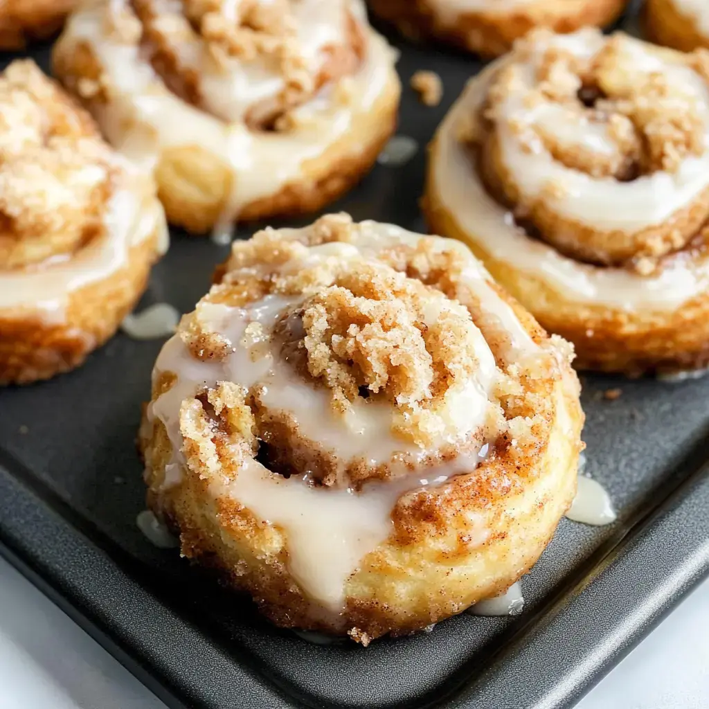 A close-up of freshly baked cinnamon rolls topped with icing and streusel, displayed on a dark baking tray.