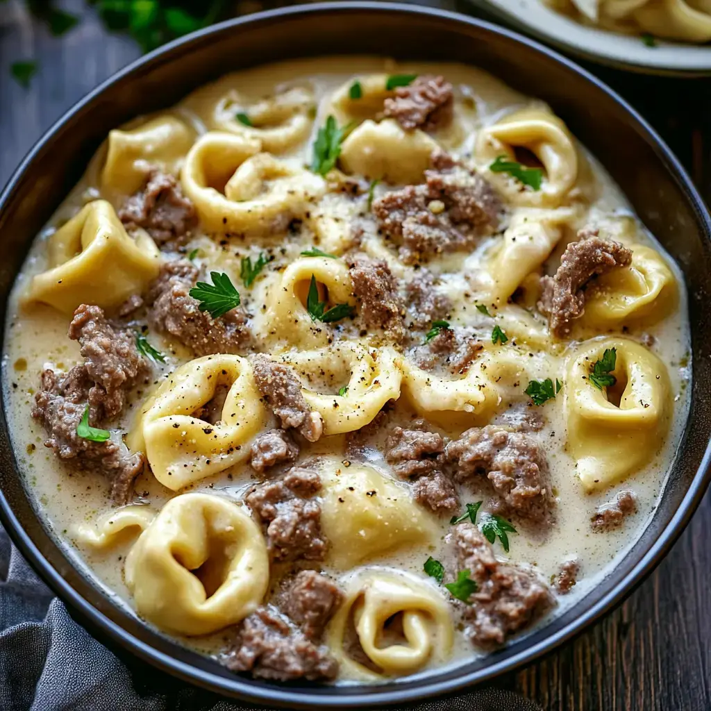 A close-up of a bowl filled with creamy tortellini and ground meat, garnished with parsley and black pepper.