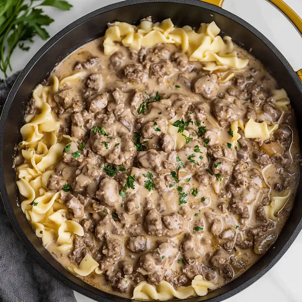 A close-up view of a skillet filled with creamy beef stroganoff served over egg noodles and garnished with chopped parsley.