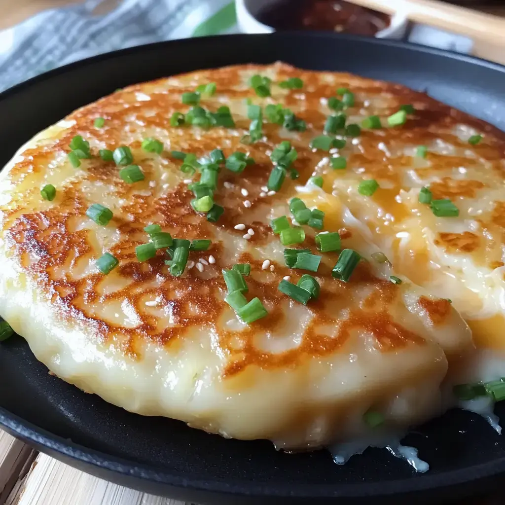 A browned, cheesy pancake is garnished with chopped green onions and served in a black skillet.