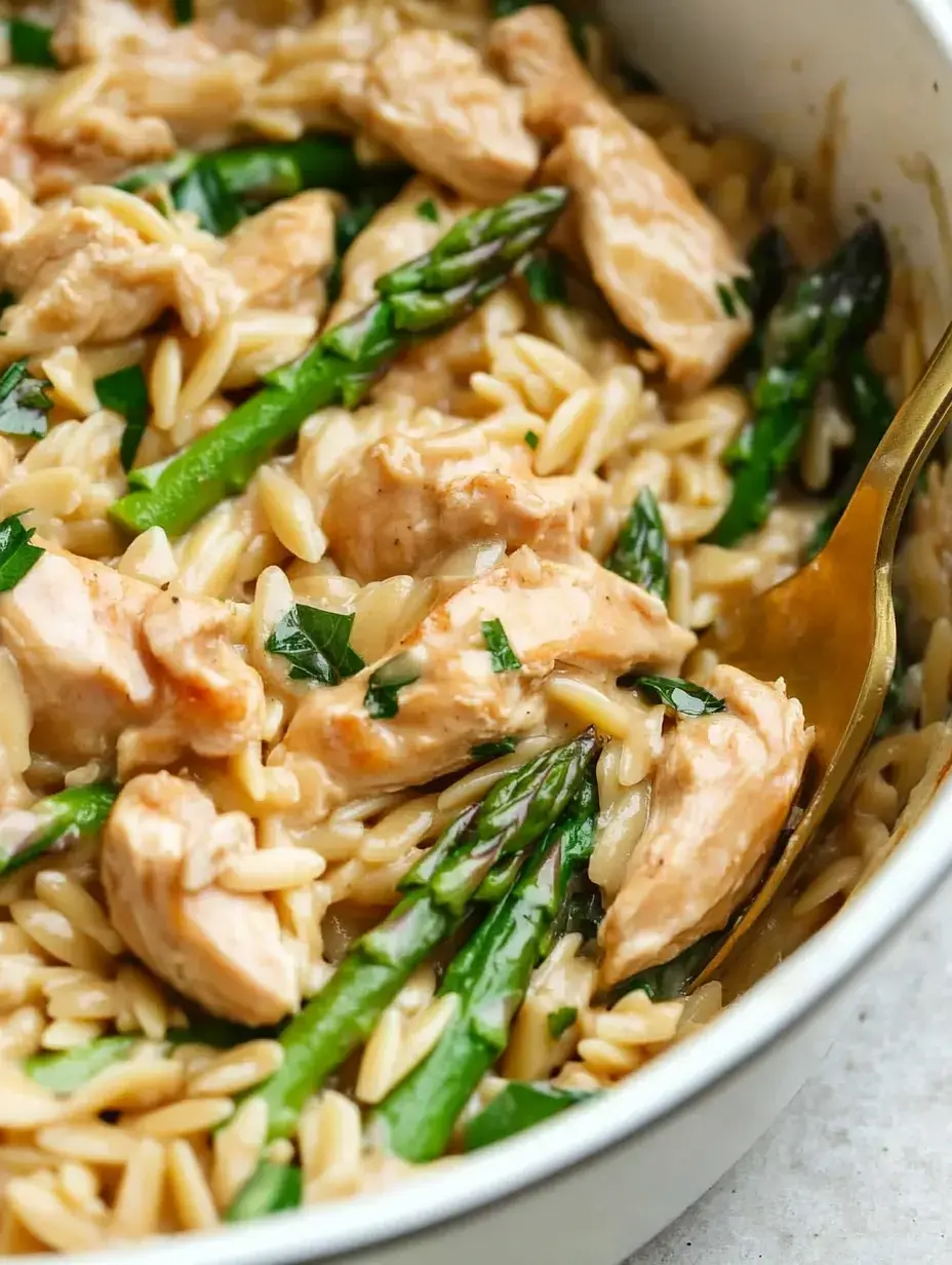 Creamy orzo pasta with chicken pieces and asparagus, garnished with fresh herbs, served in a white dish.