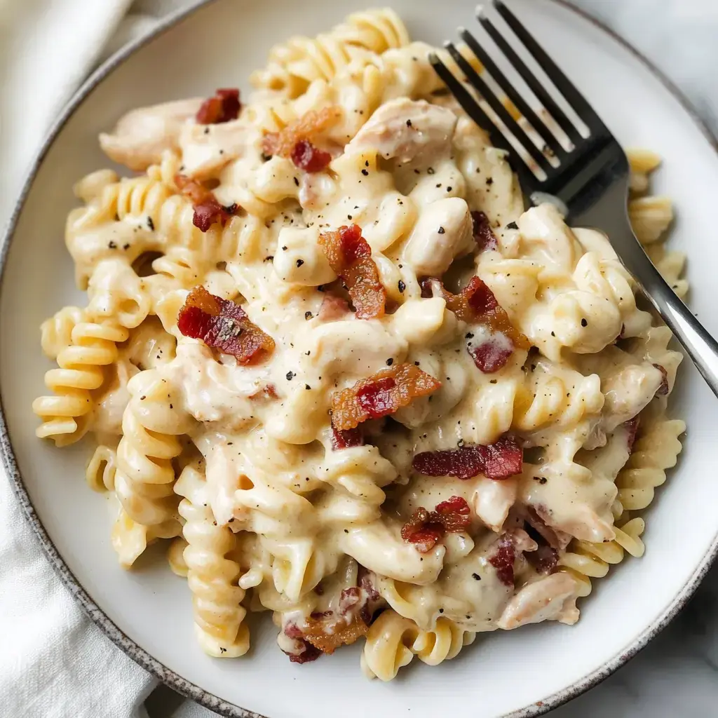 A bowl of creamy pasta with rotini, chicken pieces, and crispy bacon bits, garnished with black pepper.