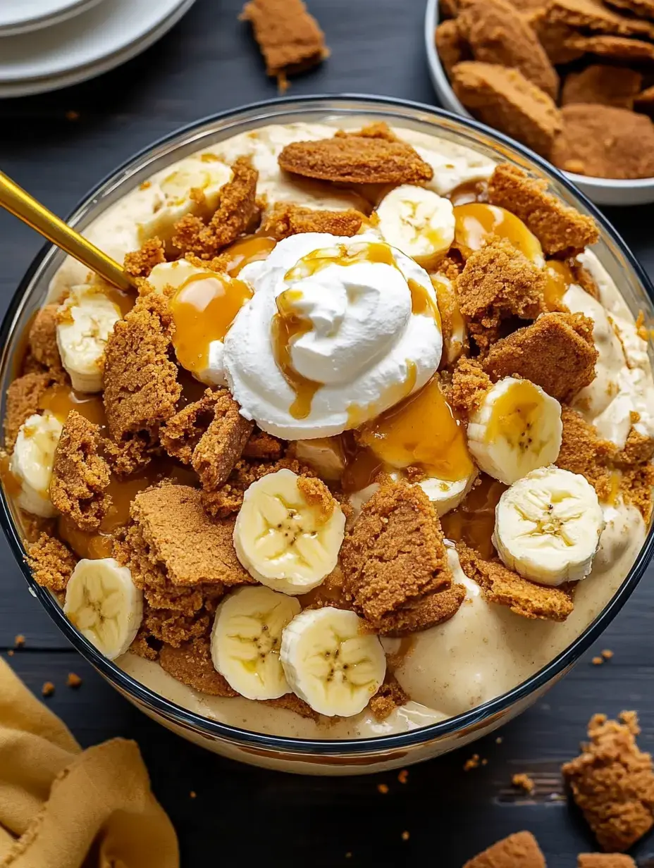 A glass bowl filled with layered dessert topped with banana slices, crushed cookies, whipped cream, and caramel sauce.