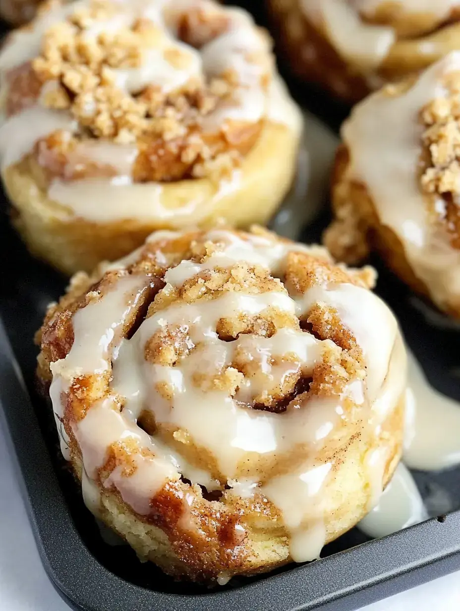 Freshly baked cinnamon rolls topped with creamy icing and crumbled topping are arranged in a dark baking tray.