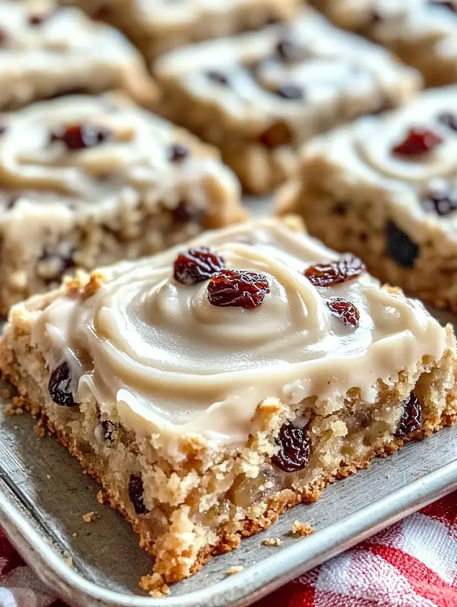 A close-up of a slice of cake topped with a smooth icing and scattered raisins, set on a tray with more cake pieces in the background.