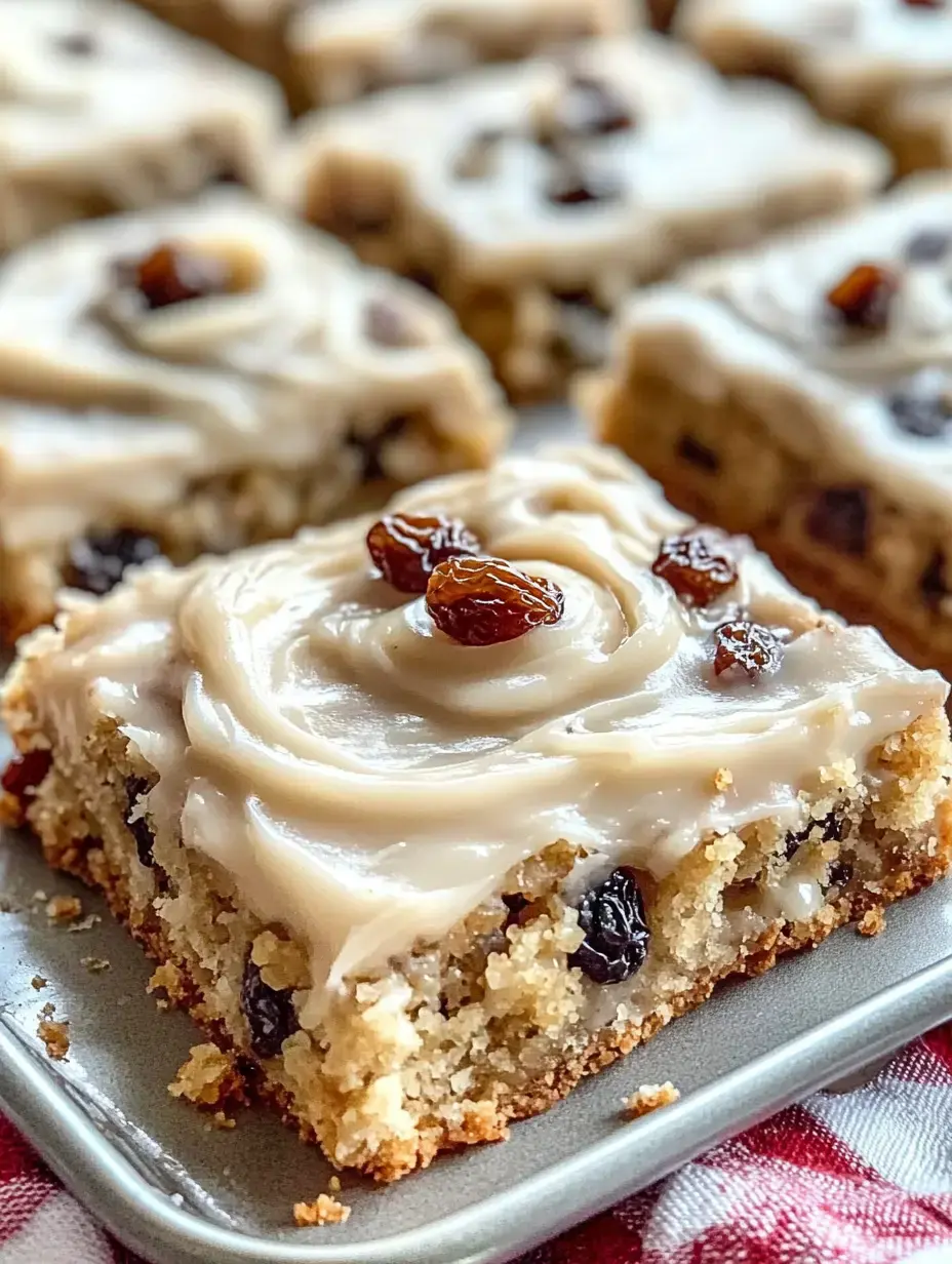 A close-up of a square piece of frosted cake topped with raisins, set on a plate with more cake pieces in the background.