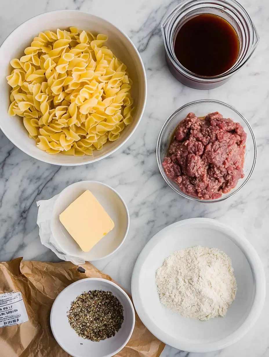 A flat lay of ingredients for a recipe, including bowtie pasta, ground beef, butter, flour, broth, and a spice mix, arranged on a marble countertop.