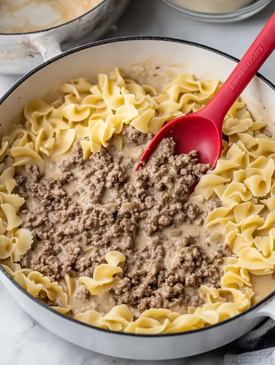 A skillet filled with cooked egg noodles and ground beef in a creamy sauce, with a red spatula mixing the ingredients.