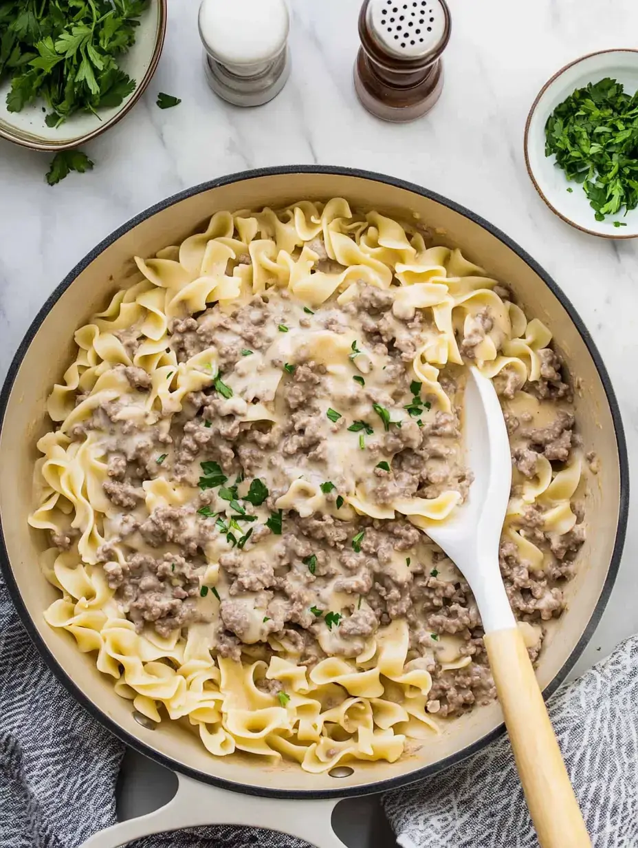 A creamy beef and noodle dish garnished with parsley, served in a large skillet alongside fresh herbs and condiments.