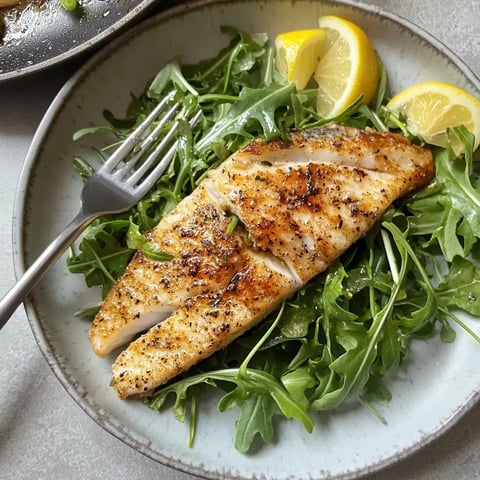 A beautifully plated piece of seared fish rests on a bed of fresh arugula with lemon wedges on the side, complemented by a fork.