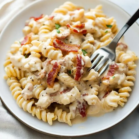 A plate of creamy pasta with bacon bits and green garnish, served with a fork.